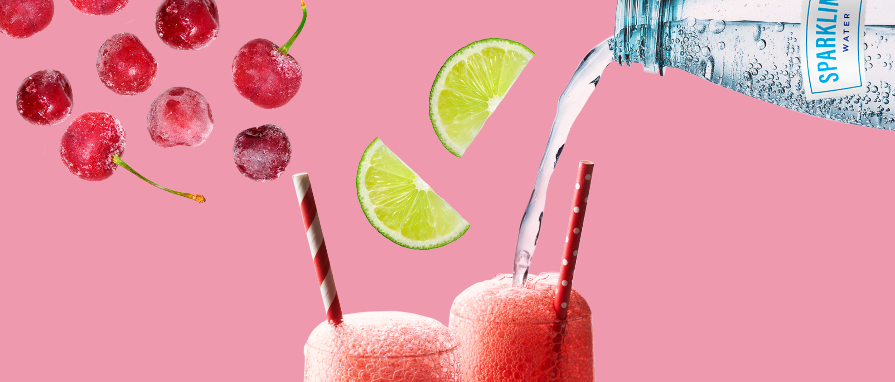 Frozen cherries, lime wedges and sparkling water float in the air over two tall glasses of a red fizzy drink.