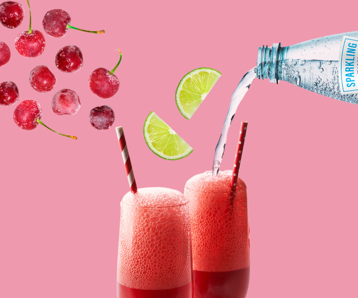 Frozen cherries, lime wedges and sparkling water float in the air over two tall glasses of a red fizzy drink