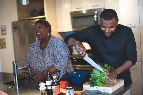 A middle-aged man with an athletic build and a middle-aged woman in a larger body prepare food in a home kitchen. The woman washes a zucchini in the sink while appearing to laugh heartily. The man is chopping a lettuce with a knife and has a smile on his face. In the foreground are various ingredients including a pepper mill, sauce bottles, baby carrots and a capsicum.