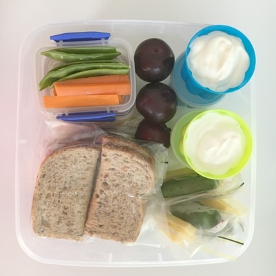A plastic box that has: wrapped up sandwiches, yoghurt in small plastic containers, vegie sticks in small containers and zip lock bags, cheese cut into cubes.
