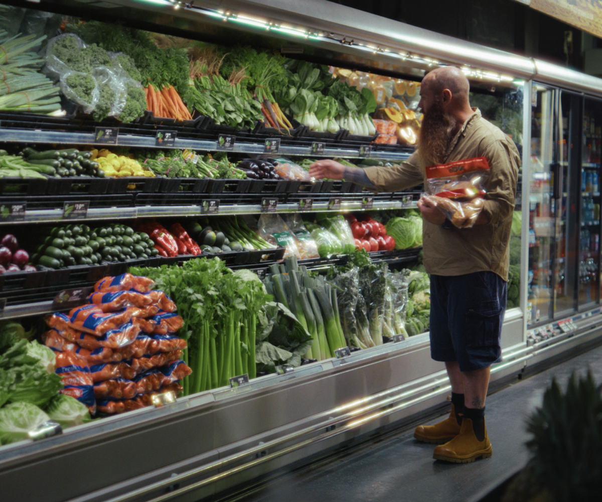 Man in work gear picks a bag of slaw of the supermarket shelf as he holds a roast chicken in his hand.