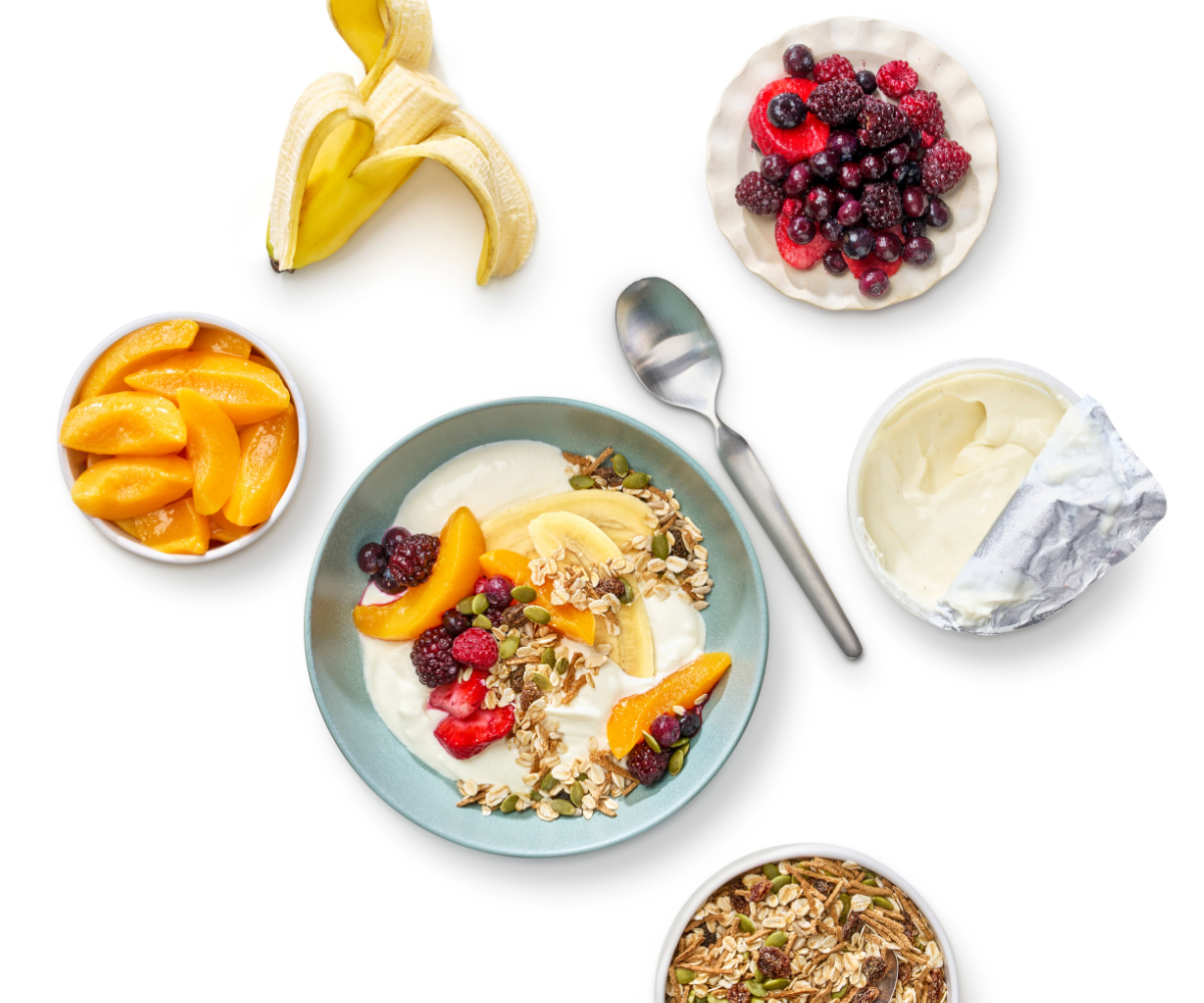 Flat lay of ingredients including yoghurt tub, opened banana, bowl of nuts and seeds, bowl of frozen berries, bowl of tinned peaches. Bowl with a combination of the ingredients is in the centre.