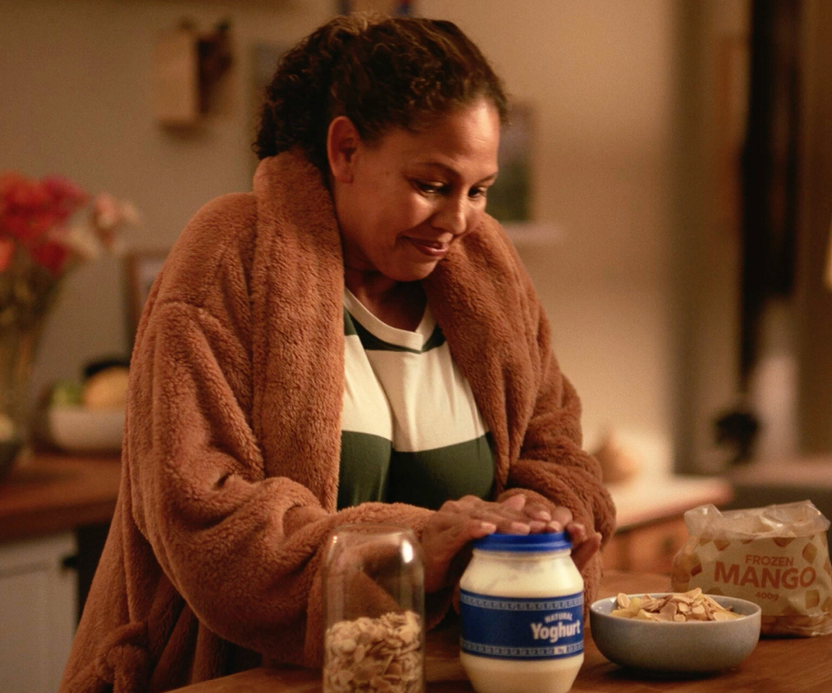 Woman smiles as she replaces the top on a yoghurt tub. A bag of frozen mangos, a jar of nuts and a bowl with a combination of the ingredients also sit on the kitchen benchtop.