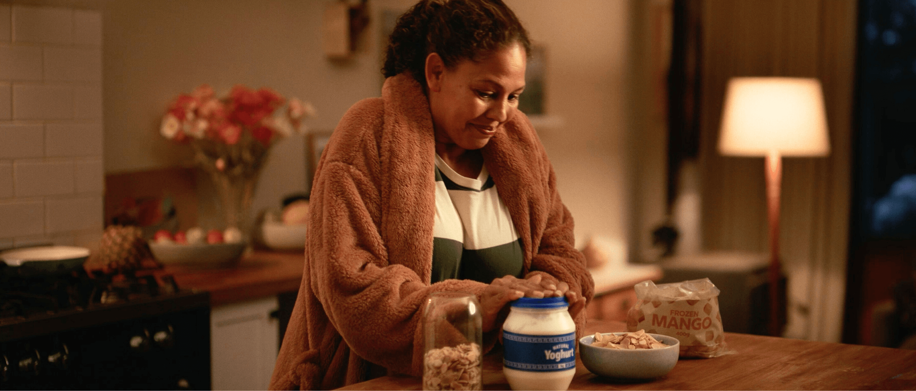 Woman smiles as she replaces the top on a yoghurt tub. A bag of frozen mangos, a jar of nuts and a bowl with a combination of the ingredients also sit on the kitchen benchtop.