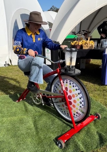 Young woman in a hat and long sleeved Cancer Council WA shirt with Aboriginal designs pedals the blender bike outside at an event. In the background a basket of bananas sits on a trestle table alongside several glasses.