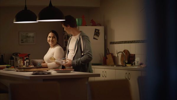 A still from the Menu App television advert. A man and woman in larger bodies are preparing a meal in the kitchen. The couple look at each other lovingly. The man holds a carton of eggs.