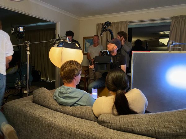 Behind the scenes shot of the filming of the Menu App television advert. A man and woman are seen from behind sitting on a couch looking at a phone that the man is holding. In the background are the film crew with various equipment including lights and a camera.