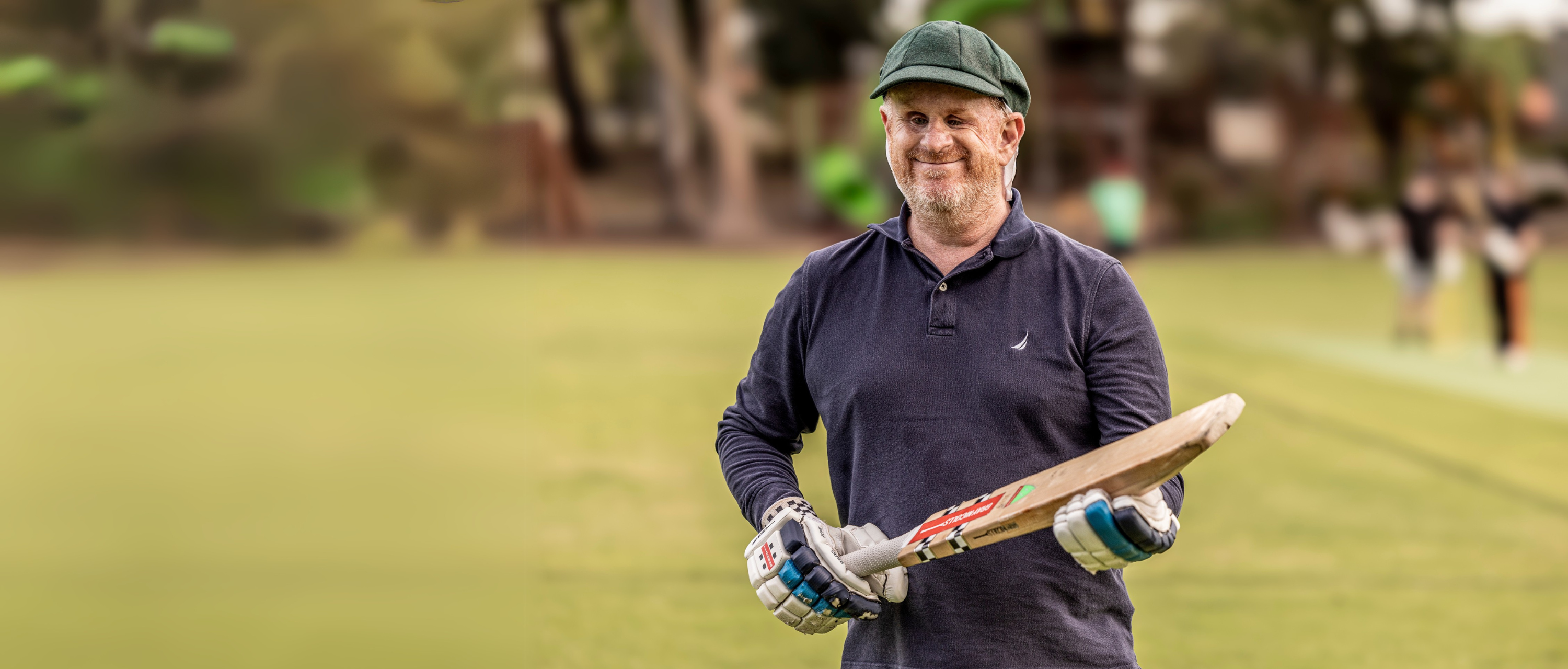 Blind cricket player stands facing camera against a blurred backdrop of a green field, he holds a cricket bat in his hands.