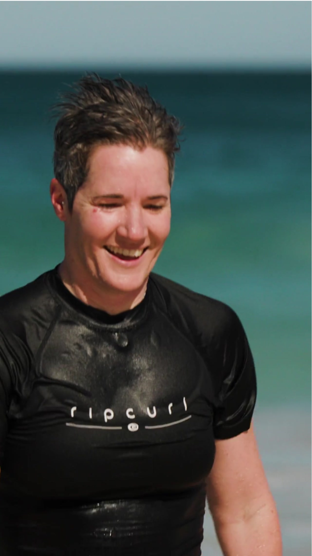 Close up of a woman with a swimming cap and goggles over the cap, beach is in the background.