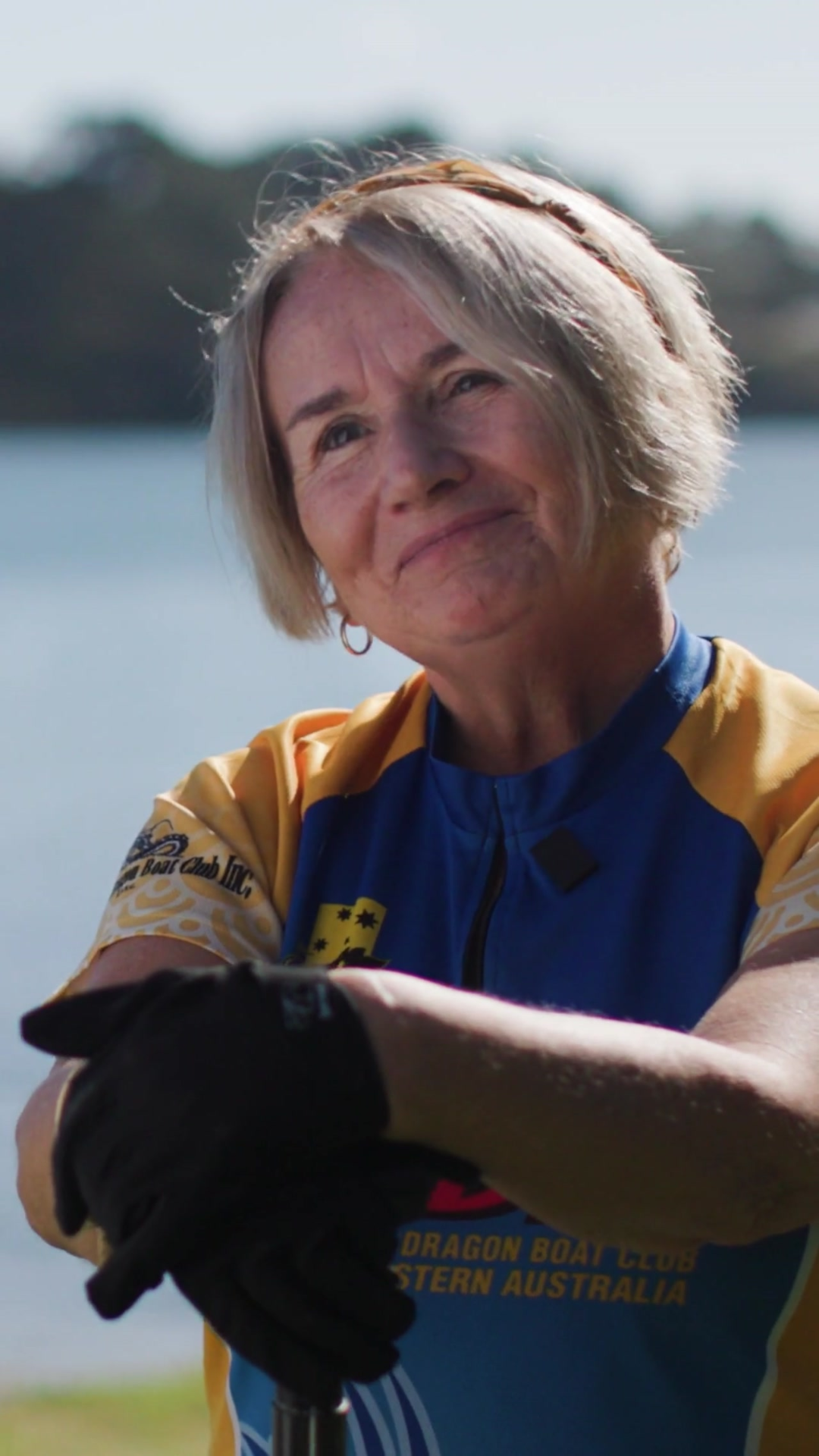 A woman smiles at the camera. The river is in the background. She rests her black gloved hands on a paddle.