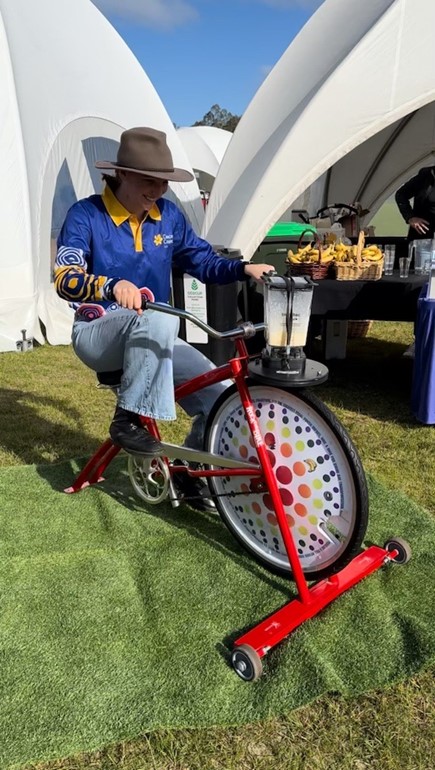 Young woman in a hat and long sleeved Cancer Council WA shirt with Aboriginal designs pedals the blender bike outside at an event. In the background a basket of bananas sits on a trestle table alongside several glasses.