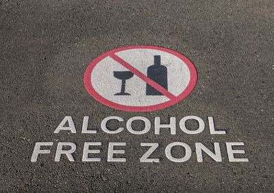 Close up of a section of bitumen road. ALCOHOL FREE ZONE is written in capital letters on the road. Icons of a wine glass and wine bottle appear in a white circle. A red outline appears around the circle and a red line crosses over the circle to indicate that these are not allowed.