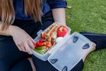 Close up of showing someone sitting on grass holding a full lunchbox of healthy food items.