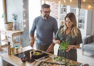 A middle-aged man and woman prepare a meal in a kitchen. The man is using a wooden spoon to stir vegetables in a frypan while looking at the woman. The woman is pulling leaves from a large bunch of herbs. In the foreground are a range of cut vegetables, some of which are on a baking tray. There are fairy lights hung behind them in the living room.