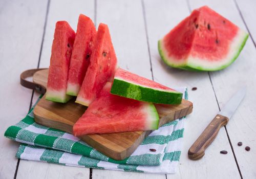 Slices of watermelon on a small wooden chopping board. A knife sits next to the board and a larger piece of watermelon is in the background.
