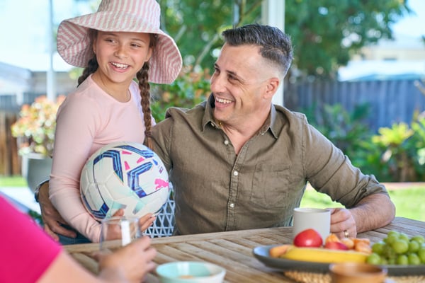 A man sits at an outdoor table in his backyard. A plate of fruit is in the centre of the table. He has one hand around a mug in front of him and one hand around his daughter who is standing next to him. The father looks towards his daughter with a smile as she looks with a smile towards her mother who is sitting out of frame. The girl is wearing a long sleeve top and broad brim hat and holds a soccer ball.