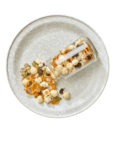 A jar spilling onto a plate. It contains popcorn, pretzels, seeds and dried fruit