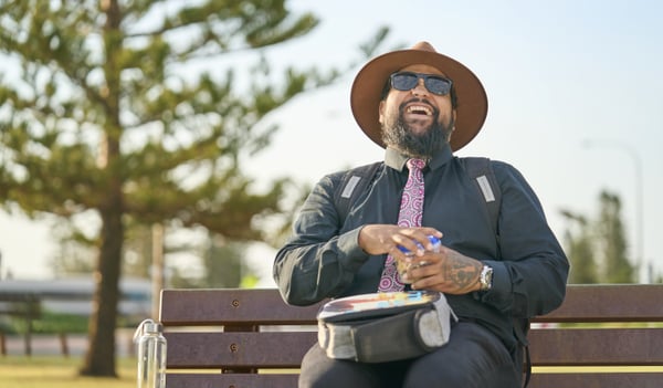 An Aboriginal man in office clothes wearing a broad brim hat and sunglasses sits on a park bench with a lunch bag on his lap and a water bottle next to him. He joyfully laughs as he opens a food container.