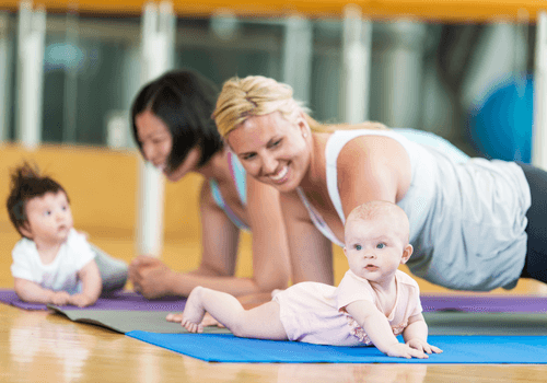 Two women do exercise on yoga mats while there babies crawl on the mats in front of them