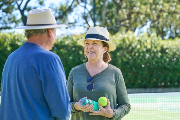 A man and woman face each other in front of the net on a tennis court. Both wear broad brim hats. The woman has a pair of sunglasses tucked into the top of her shirt and holds a tennis ball and water bottle in her hand. She is smiling towards the man.