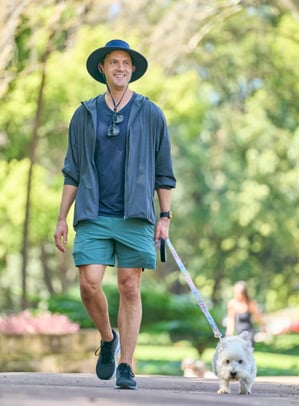 Man in a broad brim hat, long sleeves with sunglasses walks down a tree lined path holding the lead of a small white fluffy dog.