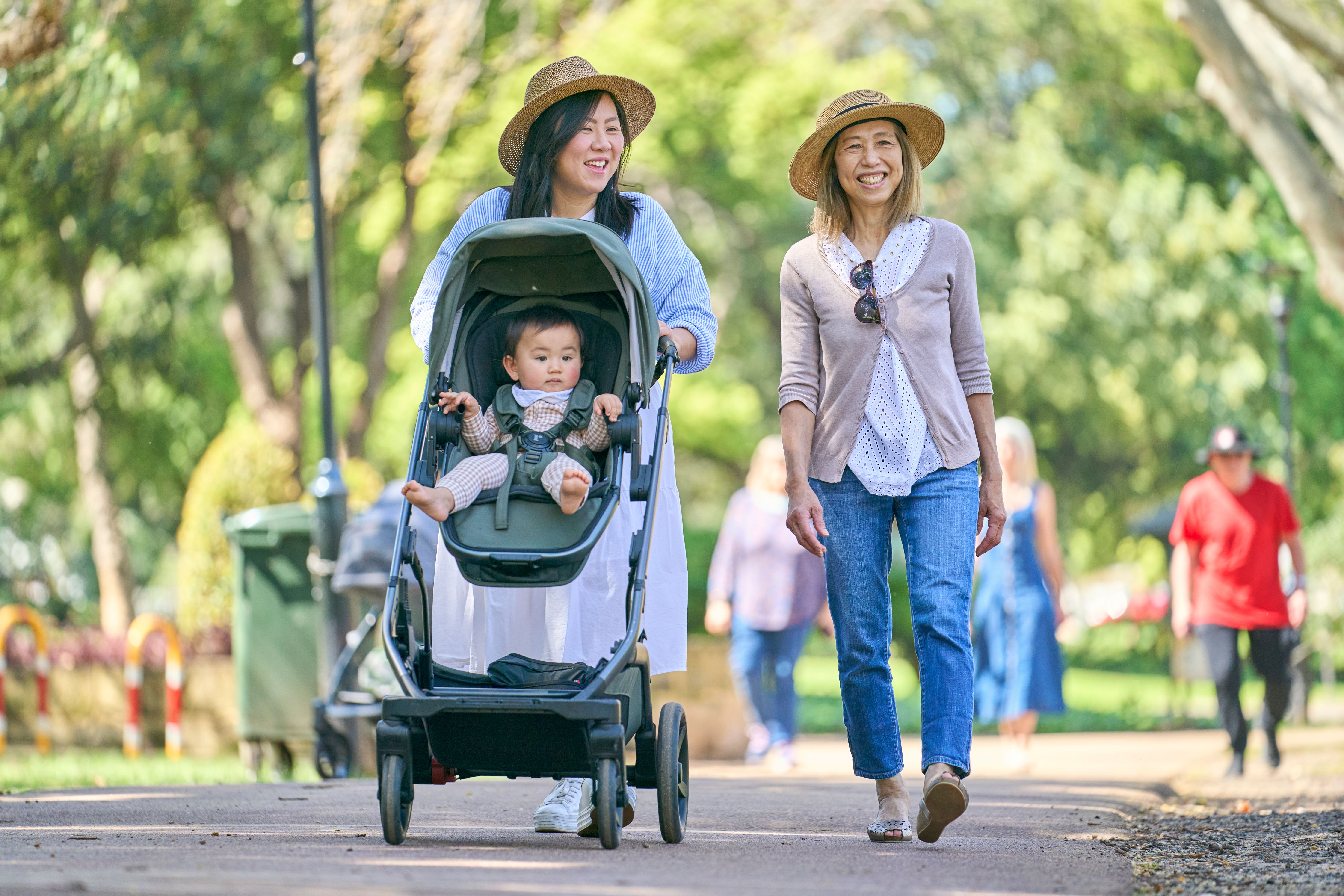 Two women walk side by side in the park while one pushes a pram. Both are smiling.