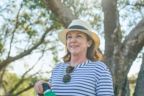 Woman holds a bottle of water. She is out in the bush with a hat, sunglasses and long sleeve top. The sun shines through the trees behind her.