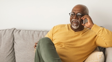 An older man with dark skin, glasses and a grey beard is smiling and talking on the phone on his couch