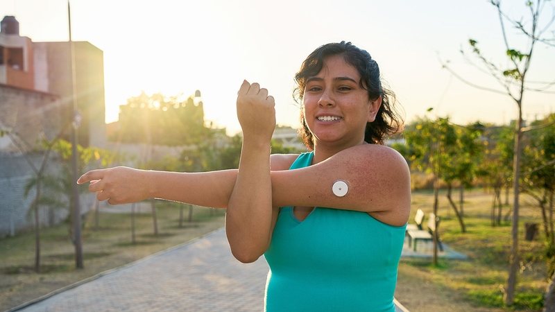 A woman is stretching outside at sunset. A continuous glucose monitor is visible on her upper arm. 