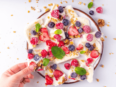 A hand reaches for a piece of yoghurt bark dotted with bright berries, crunchy nuts and fresh mint. 