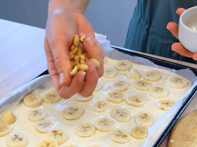 A hand sprinkles peanuts on a tray spread with yoghurt and dotted with banana slices