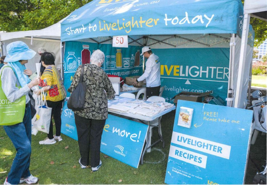A marquee with LiveLighter branding at a community event. On the table are LiveLighter branded bags. A sign next to the table encourages people to take LiveLighter recipe books. Three people are outside the stall, one of them is looking at the items on the table. A person with a LiveLighter shirt stands under the marquee facing away from the camera as they grab a box.