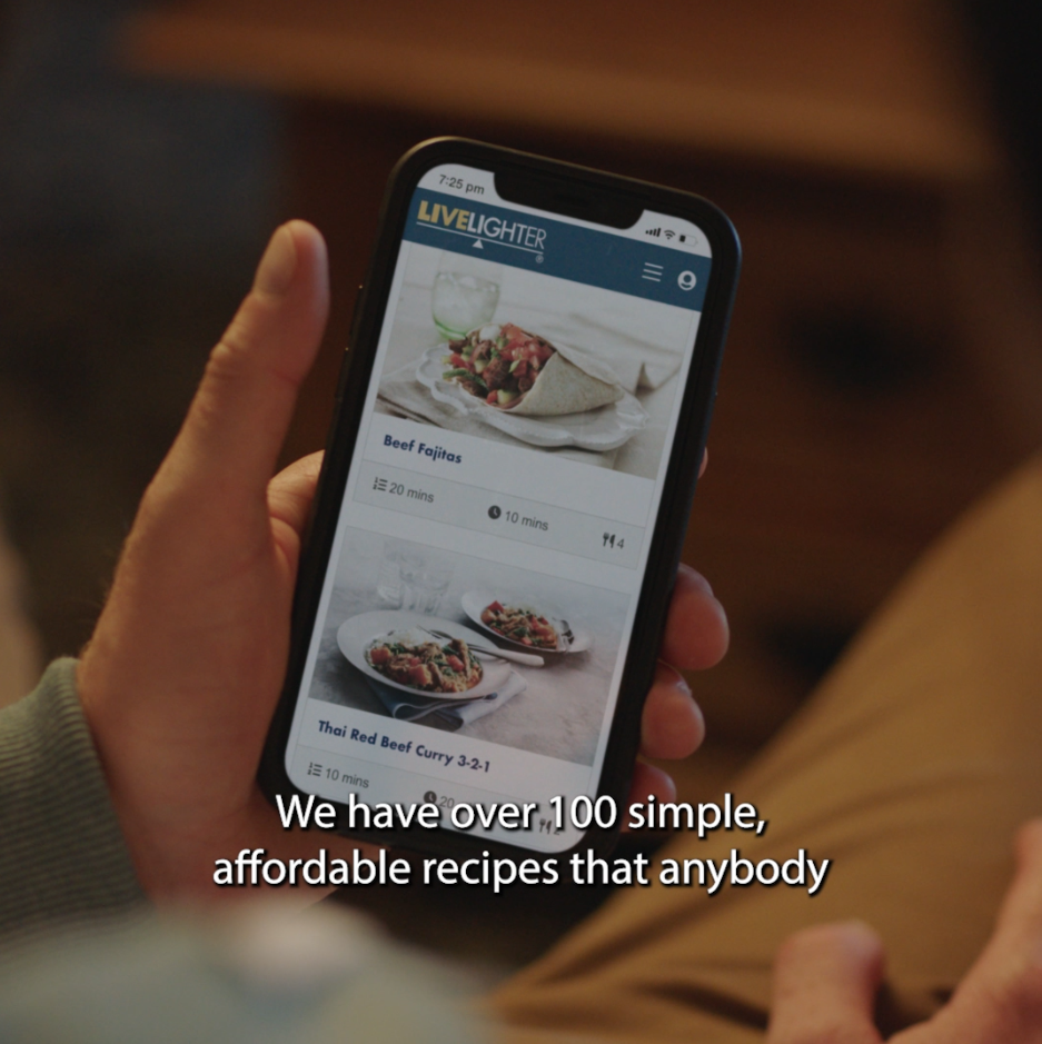 Still image from the menu app couple commercial, close up someone scrolling through their phone looking at LiveLighter recipes, close captions on the screen read 'we have over 100 simple, affordable recipes that anyone'