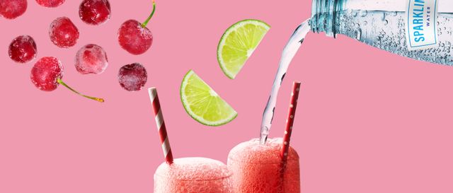 Frozen cherries, lime wedges and sparkling water float in the air over two tall glasses of a red fizzy drink