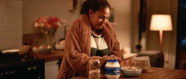 Woman smiles as she replaces the top on a yoghurt tub. A bag of frozen mangos, a jar of nuts and a bowl with a combination of the ingredients also sit on the kitchen benchtop.