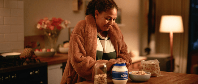 Woman smiles as she replaces the top on a yoghurt tub. A bag of frozen mangos, a jar of nuts and a bowl with a combination of the ingredients also sit on the kitchen benchtop
