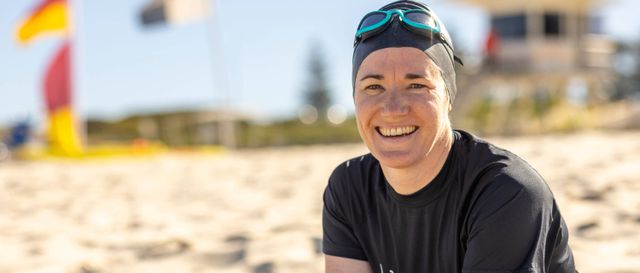 Close up of a woman with a swimming cap and goggles over the cap sitting on the beach. She is smiling and looking towards the camera.