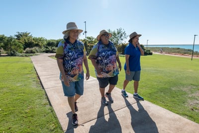 Three older women walking along the coast in Broome. They are wearing hats and colourful shirts with Aboriginal art on them. They are smiling and laughing. 