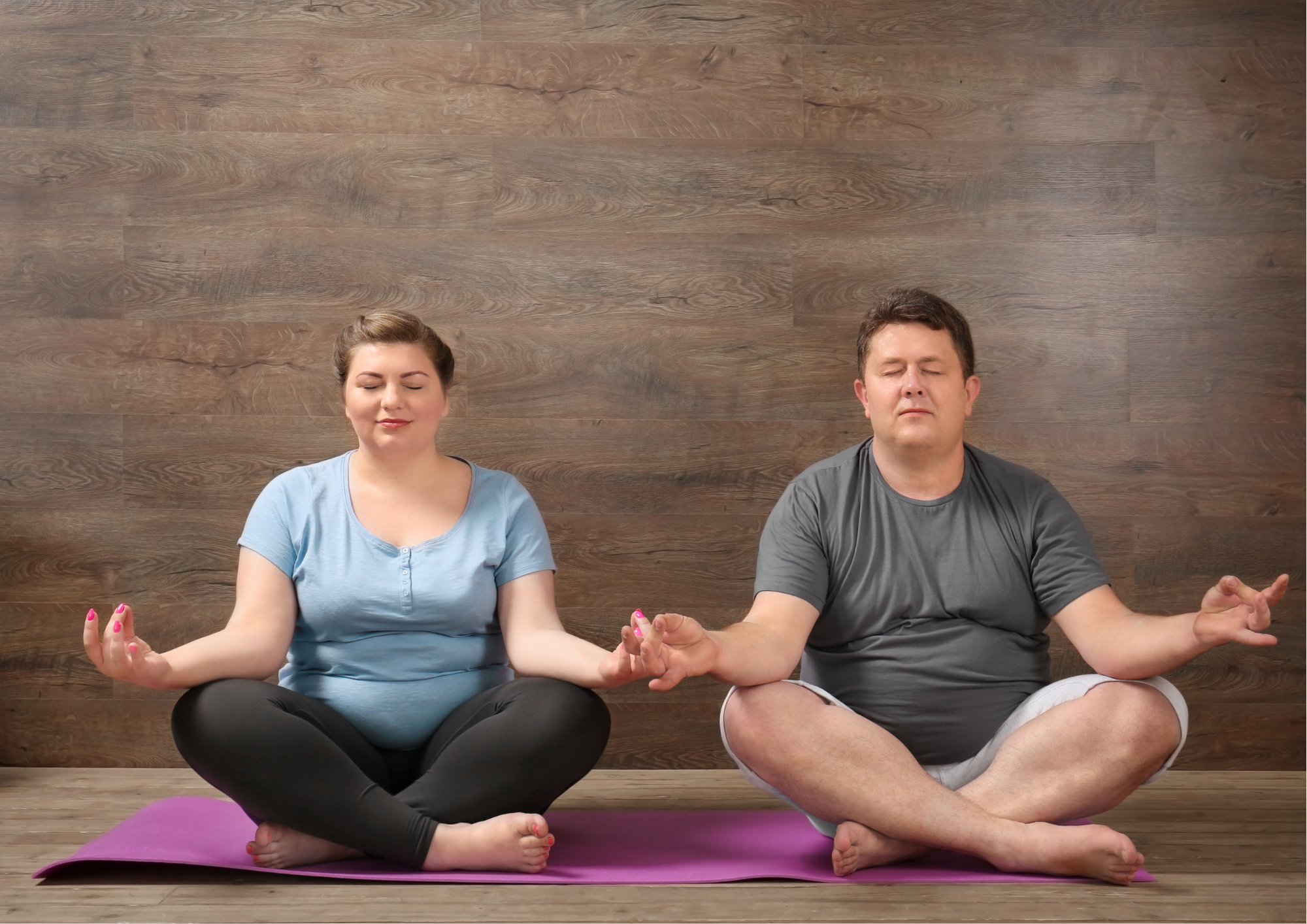a young woman and a middle aged man sit in lotus position on a yoga mat with their eyes close.