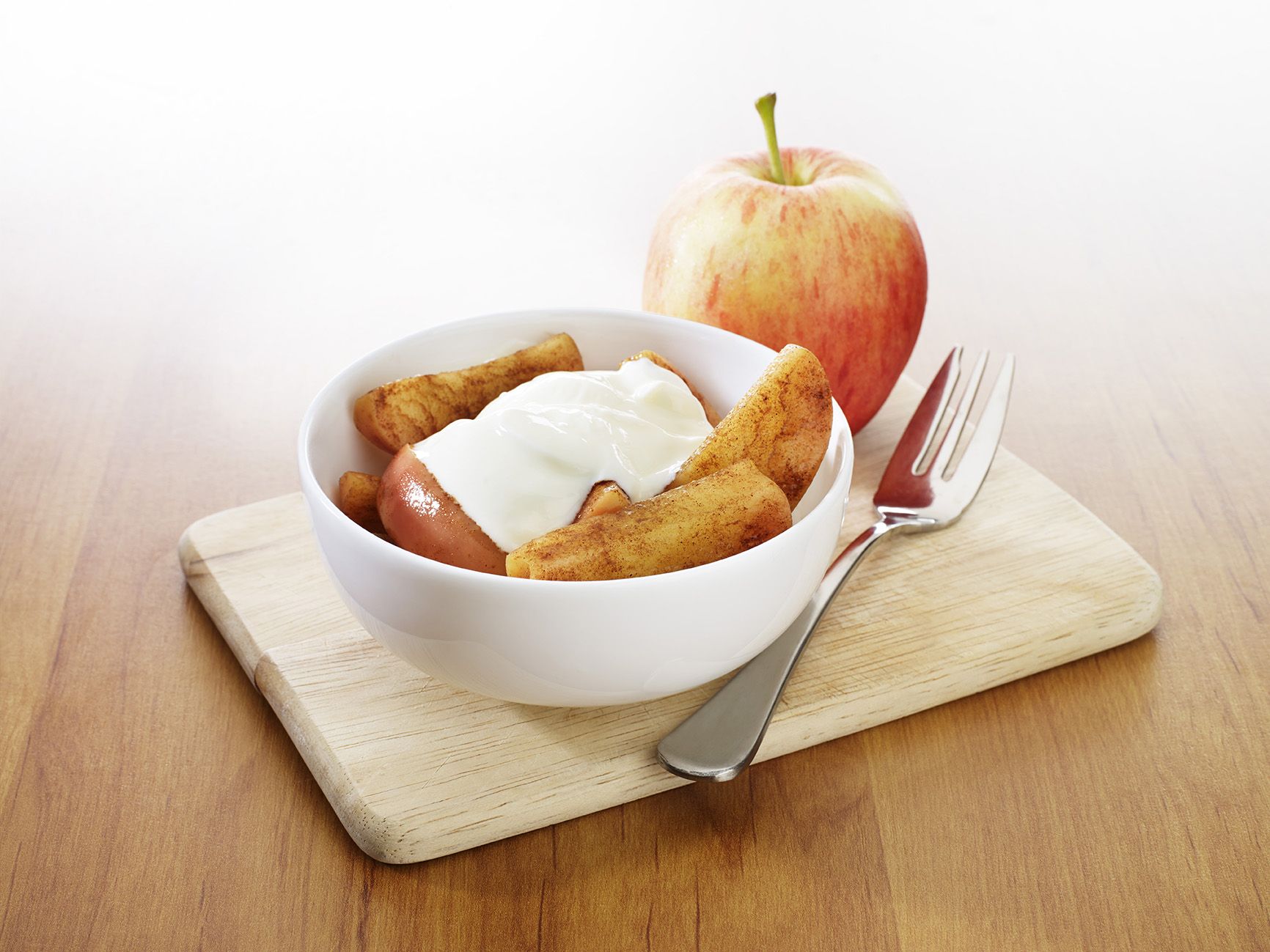 A white ceramic bowl containing sliced apples covered in cinnamon topped with a dollop of yoghurt. A whole apple sits in the background.