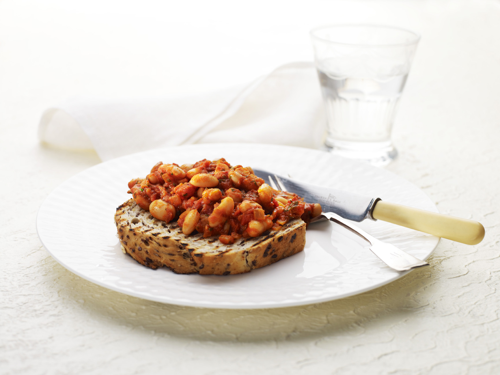 A close up of a white china plate with a slice of multigrain toast, topped with a tomato bean mix. A knife and fork are on the plate. In the background is an out of focus white tea towel and a glass of water.