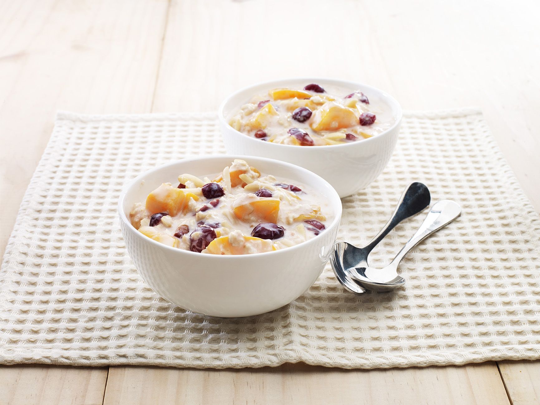 Two white ceramic bowls containing a wet mixture of oats, peach slices and cranberries.