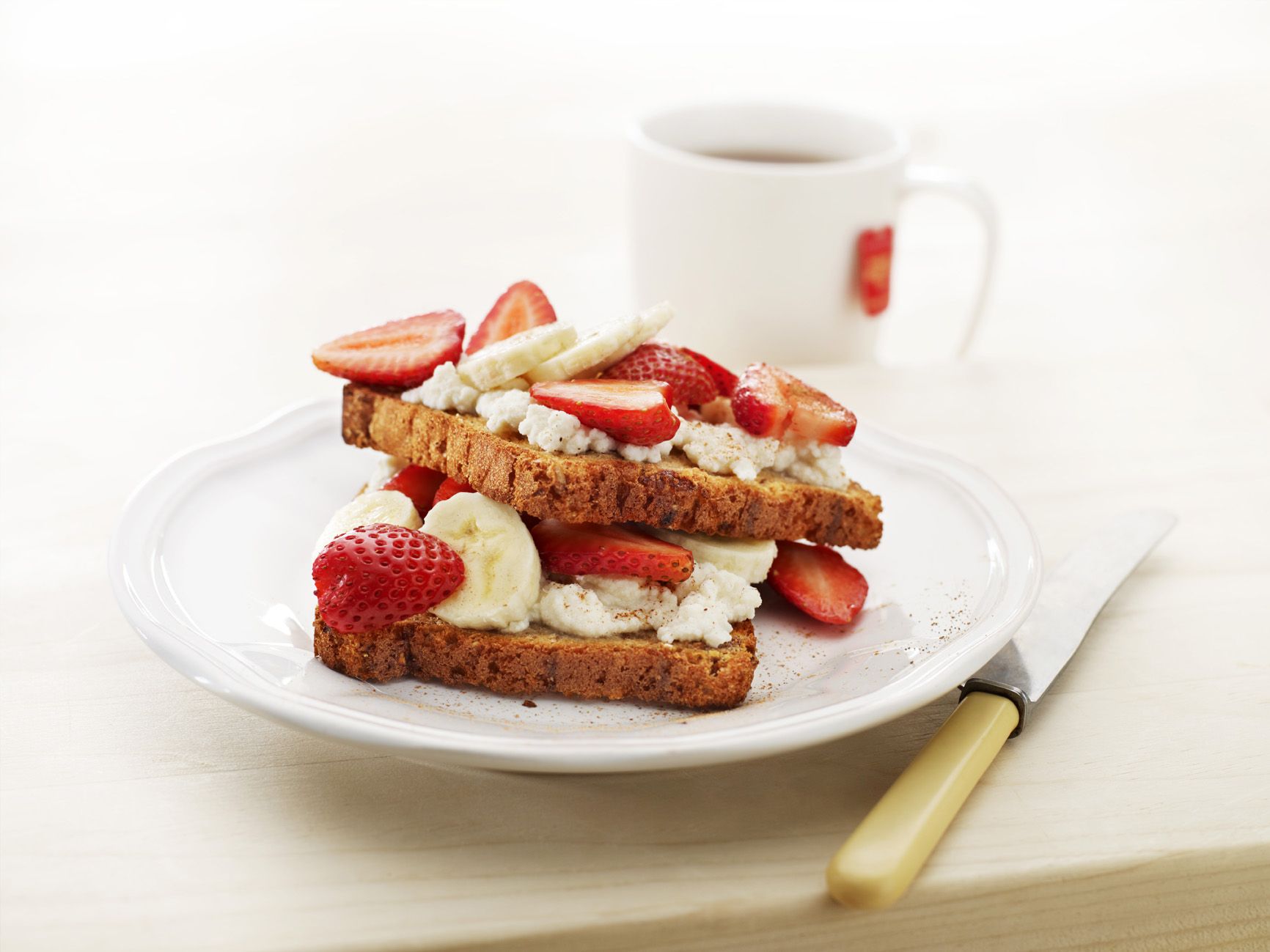 A white ceramic plate with two slices of bread topped with ricotta, sliced banana and sliced strawberries. A cup of black tea sits in the background with the teabag still in it.