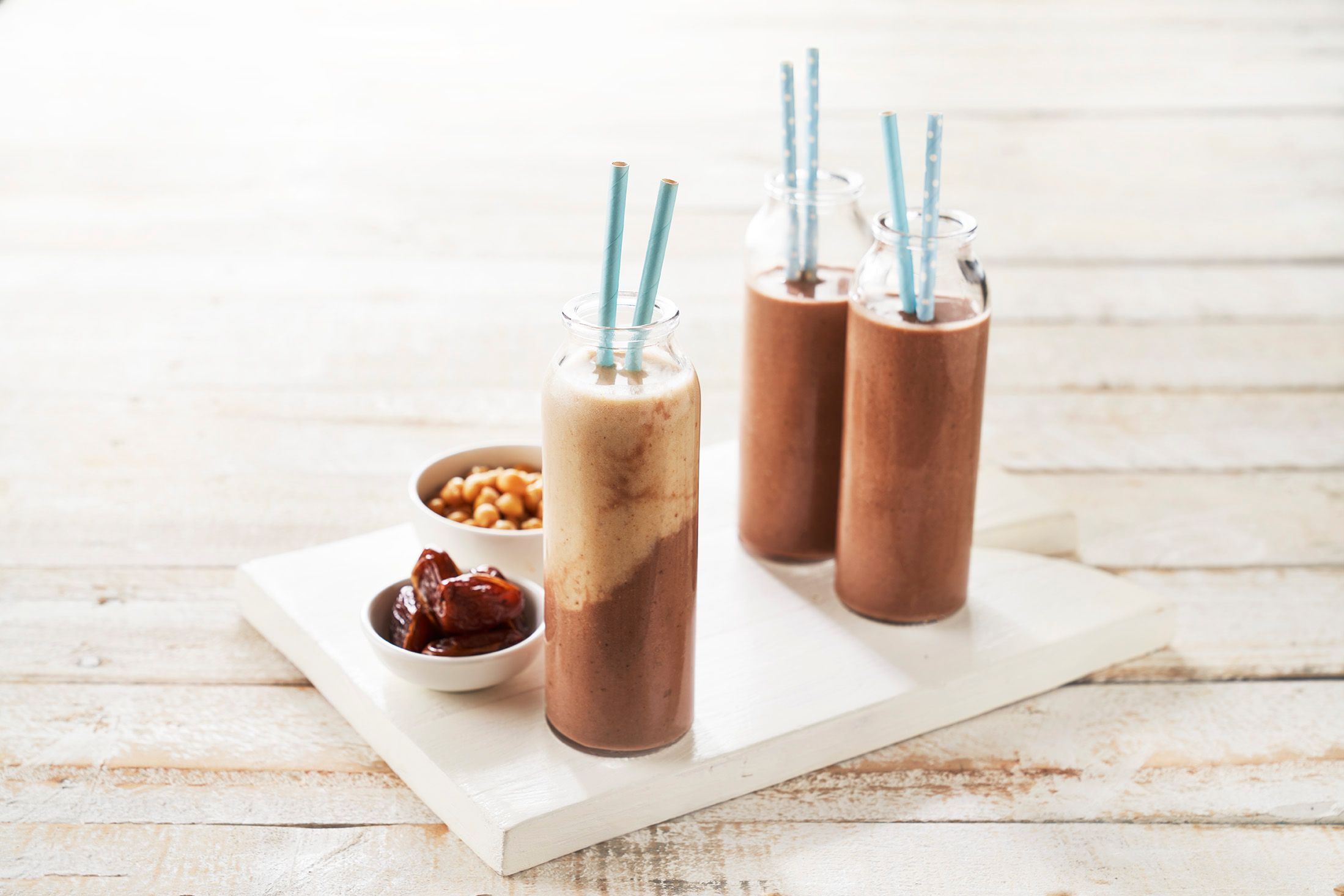 Three tall glass jars containing a creamy chocolate coloured liquid. Each glass contains two light blue paper straws. Small ramekins sit next to the glasses, one contains dates and the other contains chickpeas.