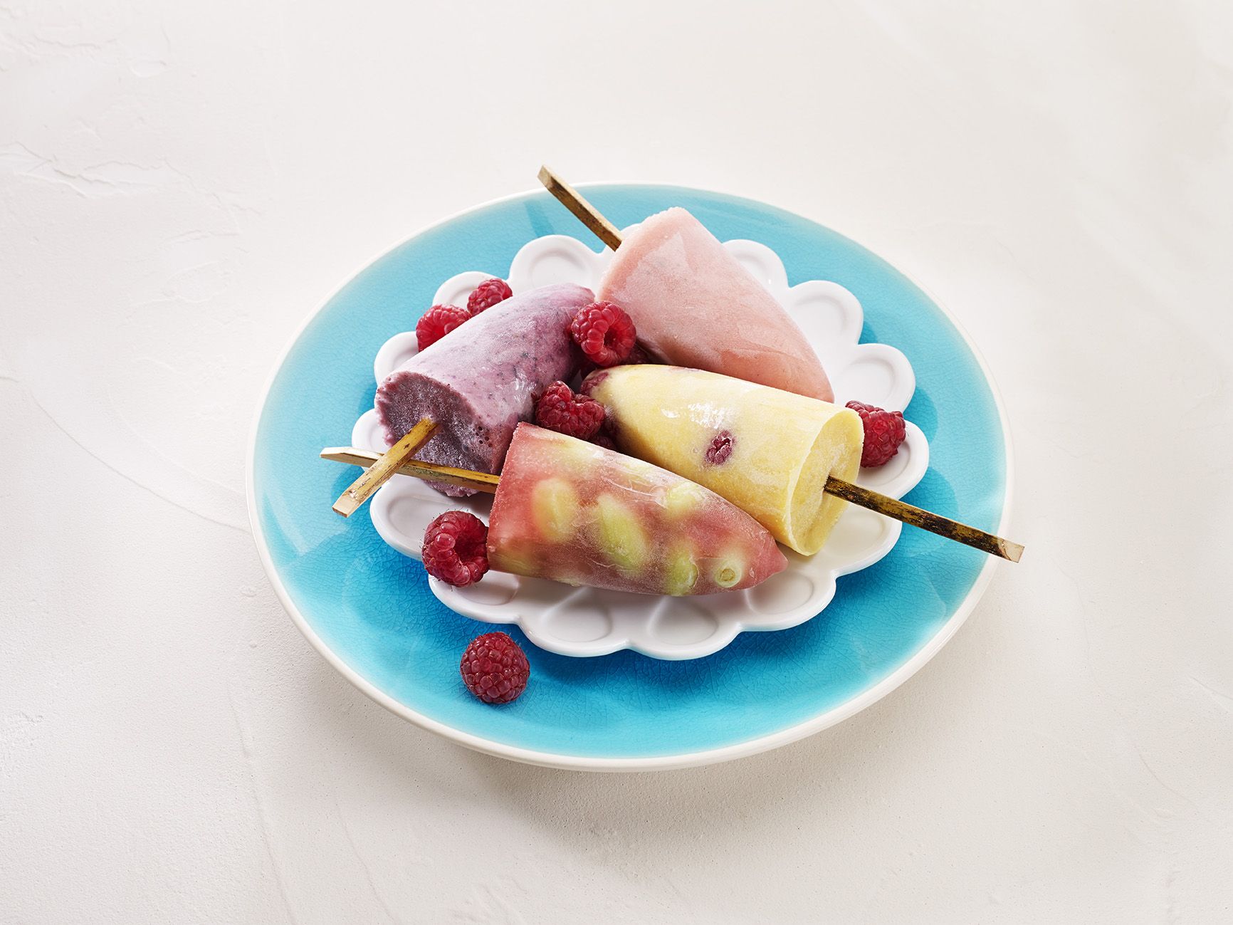 Four icepoles sitting on a light blue plate. Each icypole has a different colour. A few raspberries are scattered on the plate. 