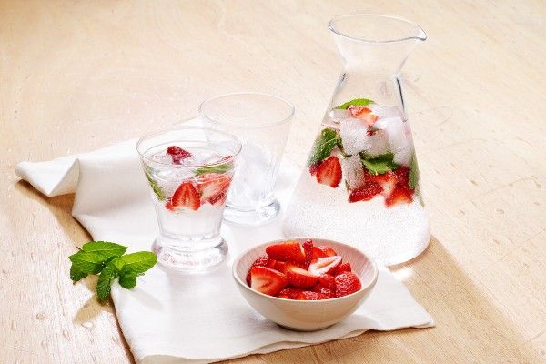  A glass jug with two glasses containing sliced strawberries and mint with fizzy water